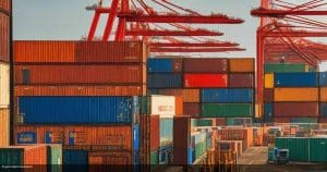 Stacked shipping containers in red, blue and green beneath red gantry cranes at a container port in evening light